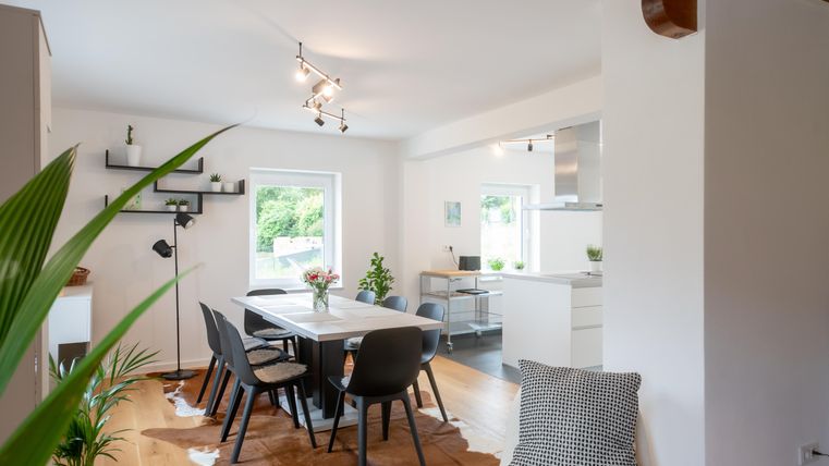 Ein modernes Wohnzimmer mit einem Esstisch und schwarzen Stühlen. Große Fenster lassen viel Tageslicht herein und Pflanzen sorgen für eine gemütliche Atmosphäre.