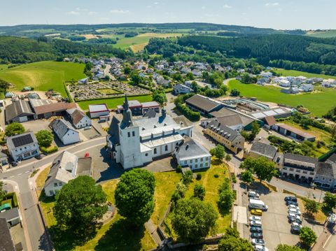Eine malerische Landschaft mit einem weißen Kirchengebäude in der Mitte. Im Hintergrund sind grüne Felder und kleine Häuser zu sehen.