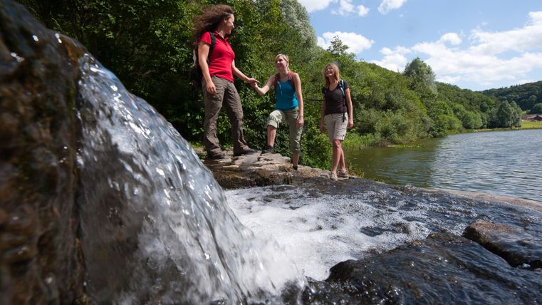 Three women hike along a waterfall in a wooded area.