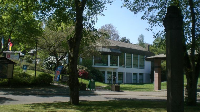 Een modern gebouw omringd door bomen en groenvoorzieningen. De lucht is blauw en het is mooi zonnig.