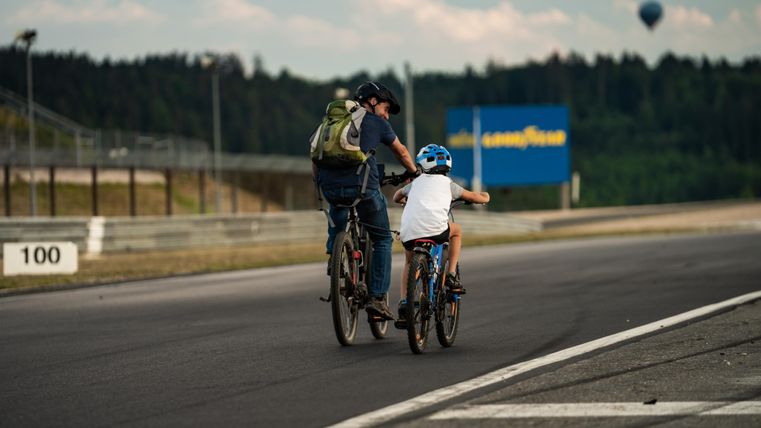 Ein Vater fährt mit seinem Kind auf Fahrrädern. Im Hintergrund sind eine Naturkulisse und ein Heißluftballon zu sehen.
