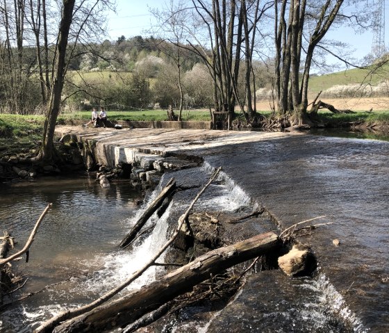 Zwei Personen sitzen auf einer Brücke über der Nims in einer grünen, ländlichen Umgebung. Der Fluss fließt ruhig unter der Brücke hindurch., © Tourist-Information Bitburger Land_Uschi Hallet