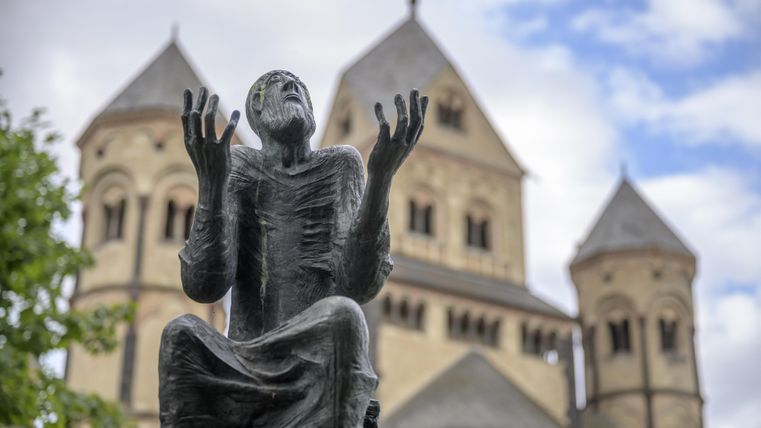 Eine Statue eines betenden Mannes steht vor einer alten Kirche. Die Wolken sind teilweise sichtbar und verleihen der Szene eine nachdenkliche Stimmung.