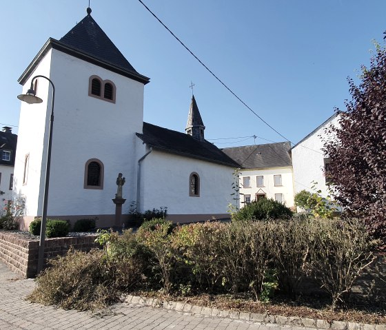 La chapelle Sainte-Lucie se trouve dans un village, entourée de maisons et d'arbres, sous un ciel bleu clair., © TI Bitburger Land, Steffi Wagner