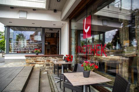 Ein einladendes Café mit einer großen Fensterfront und blühenden Pflanzen vor der Tür. Die moderne Architektur und das gemütliche Ambiente laden zum Verweilen ein.