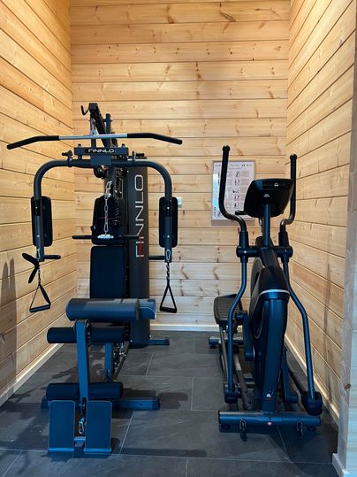 Ein kleiner Fitnessbereich mit verschiedenen Trainingsgeräten. Die Wände sind aus Holz und der Boden ist gefliest.