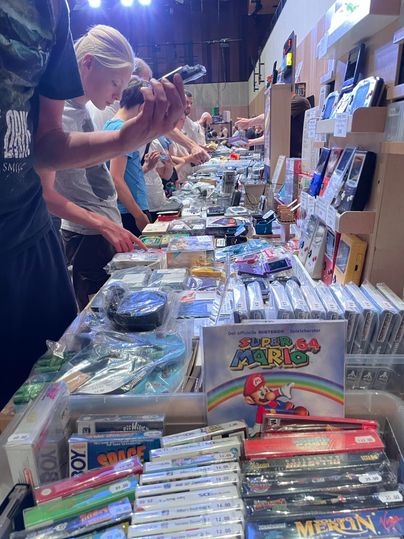 Ein belebter Flohmarkt mit Tischen voller Videospiele und Konsolen. Viele Käufer durchstöbern die angebotenen Produkte.