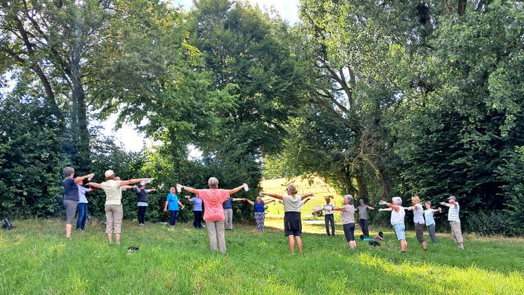 Eine Gruppe von Menschen macht Übungen im Freien. Sie stehen auf einer Wiese umgeben von Bäumen.