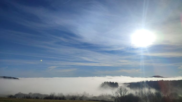 A vast landscape shrouded in thick fog and bathed in bright sunlight. The sky is dotted with soft blue and white clouds.