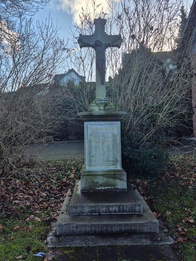 Une croix en pierre sur un socle, entourée de buissons secs et de feuilles fanées. Le ciel est partiellement nuageux.