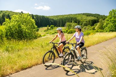 Zwei Frauen fahren Fahrrad auf einem Radweg, links und rechts ist Wiese