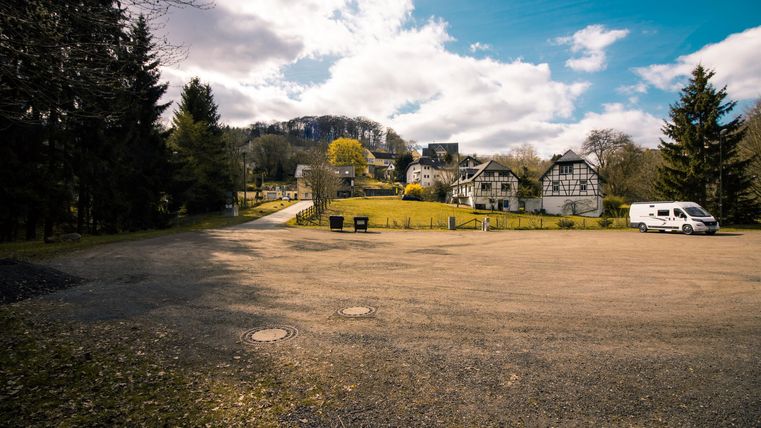 Ein großer Parkplatz mit Blick auf ein ruhiges Wohngebiet und grüne Wiesen. Im Hintergrund sind die Berge und ein blauer Himmel mit einigen Wolken sichtbar.