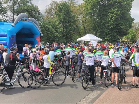 A large group of cyclists gathers in front of a colorful bounce house. The atmosphere is lively and friendly.