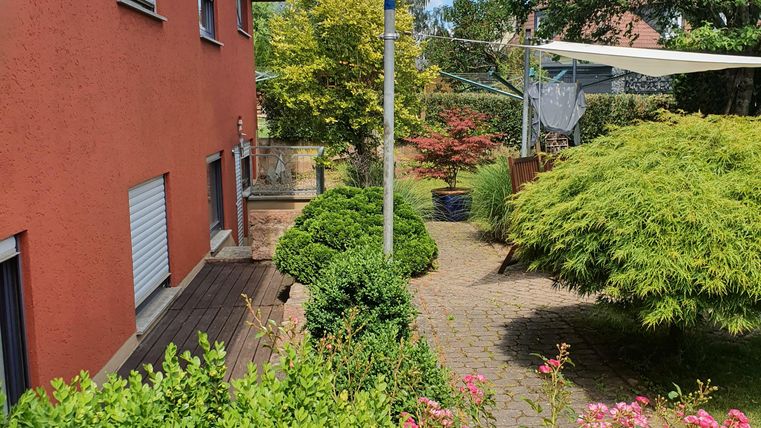 A well-kept garden with colorful flowers and a narrow path. In the background, there is a red house and a blue sky with some clouds visible.