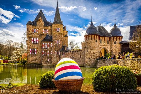 Eine malerische Burg mit einem bunten Osterei im Vordergrund. Der Himmel ist klar und die Umgebung ist grün und einladend.