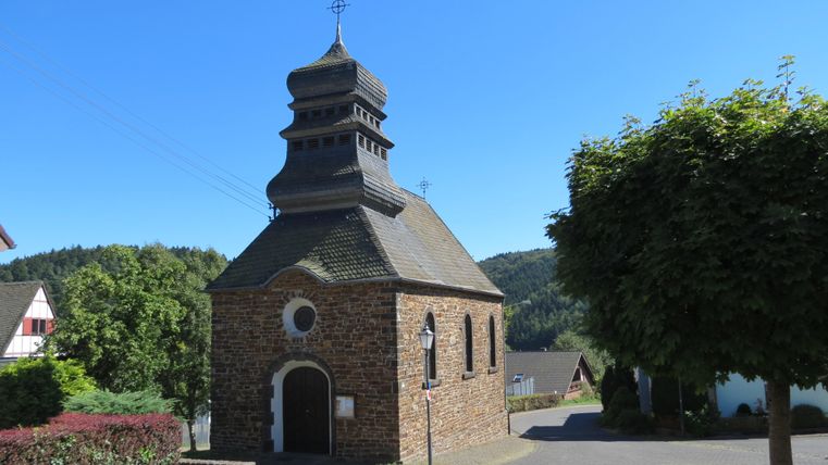 A small stone church with a distinctive roof stands on a quiet street. Surrounded by green habitat, the place exudes a peaceful atmosphere.