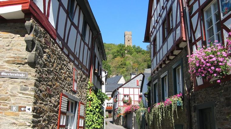 Eine malerische Gasse mit Fachwerkhäusern und blühenden Blumen. Im Hintergrund ragt ein Turm auf einem Hügel empor.