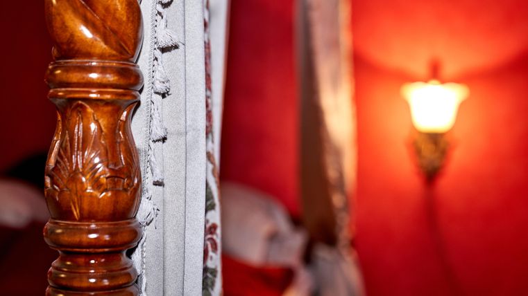 Ein detailreicher Holzpfosten eines Bettes steht im Vordergrund. Im Hintergrund sieht man eine warme Lichtquelle an einer rot gestrichenen Wand.