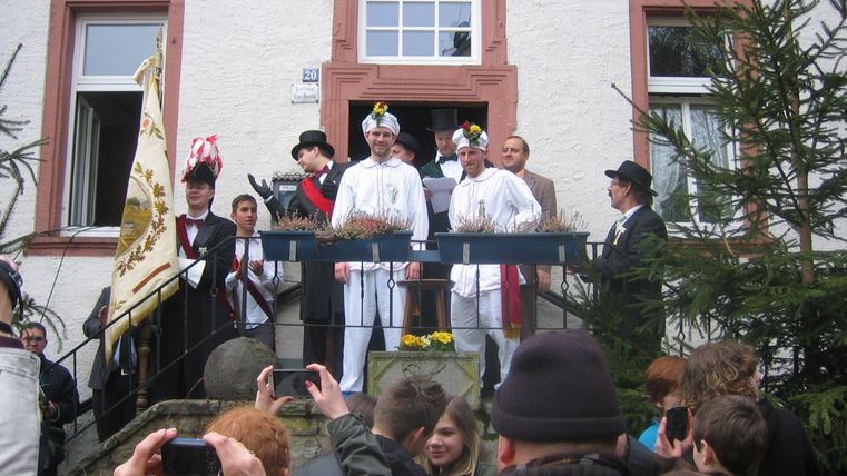 A festive gathering in front of a building with people in traditional clothing. At the center are two men who appear to be celebrating.