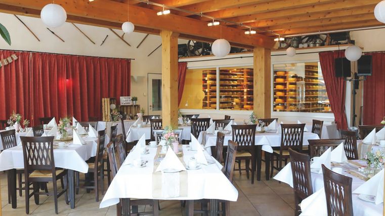 Ein Raum, der mit Tischen gefüllt ist, die Tische sind mit einer weißen Tischdecke und gefalteten Servietten gedeckt, im Hintergrund sieht man die Käserei des Gröner Hofs
