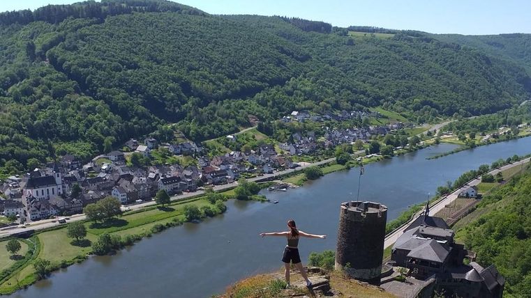 A person stands on a hill with a sweeping view of a village and the river. Surrounded by green hills, the scene offers a calm, natural beauty.