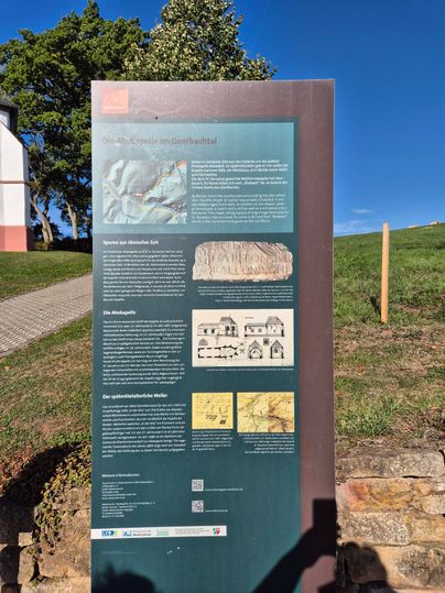 Ein Informationsschild steht auf einer Wiese, umgeben von Bäumen. Das Schild enthält Texte und Bilder zu einem historischen Thema.
