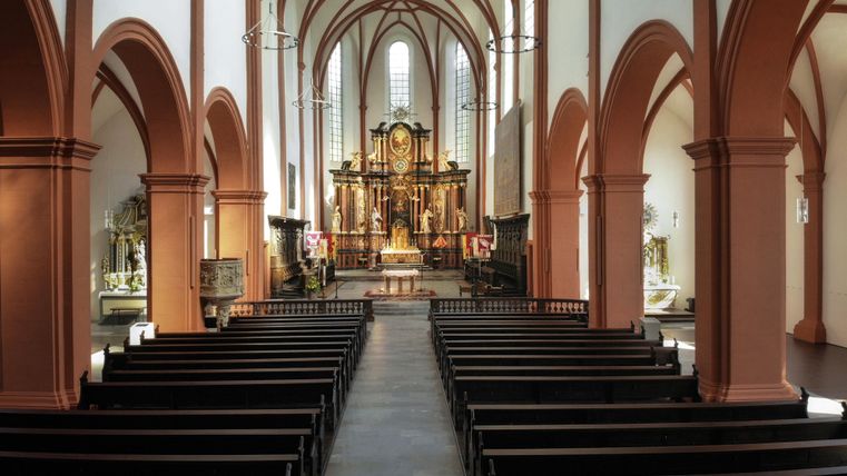 Eine helle Kirche mit beeindruckenden Bögen und einer detailreichen Altaranlage. Die Bänke sind ordentlich aufgereiht und laden zum Verweilen ein.