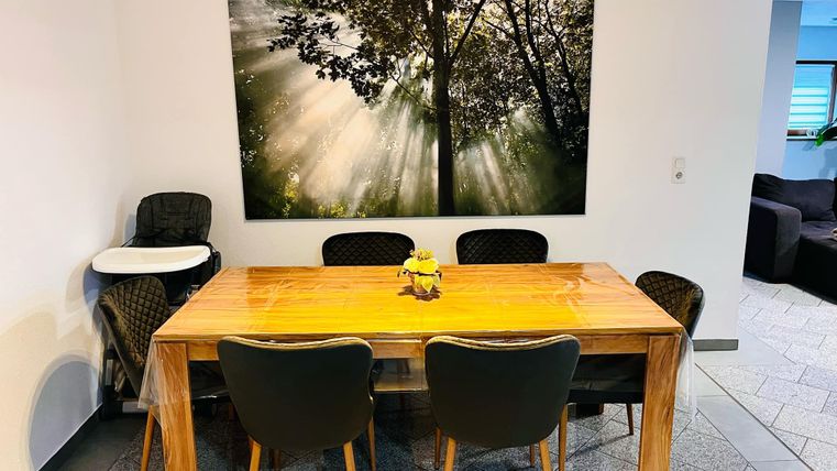 Een gezellige eetruimte met een houten tafel en zwarte stoelen. Aan de muur hangt een groot schilderij van bomen met zonnestralen.
