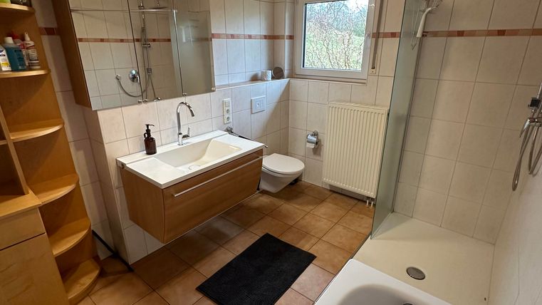 Ein modernes Badezimmer mit einer Badewanne und einer Dusche. Es gibt Holzelemente und eine helle Atmosphäre durch das Fenster.