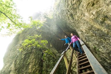 Zwei Personen stehen auf einer Treppe die in  die Buchenlochhöle führt. Links und rechts der Personen ist der Fels zusehen.
