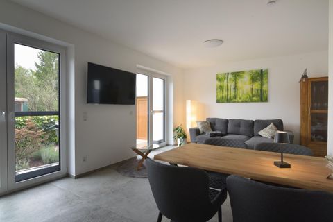 Ein modernes Wohnzimmer mit einem grauen Sofa und einem Holztisch. Große Fenster lassen viel Licht herein und ein Kunstwerk mit grünen Bäumen schmückt die Wand.