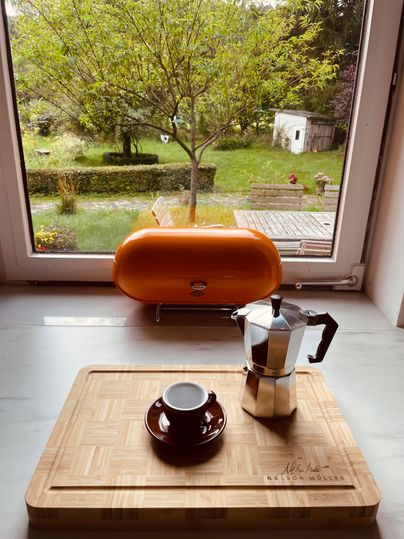 Une scène de cuisine douillette avec une cafetière moka et une tasse sur une planche en bois. En arrière-plan, le jardin avec un arbre et des meubles est visible.
