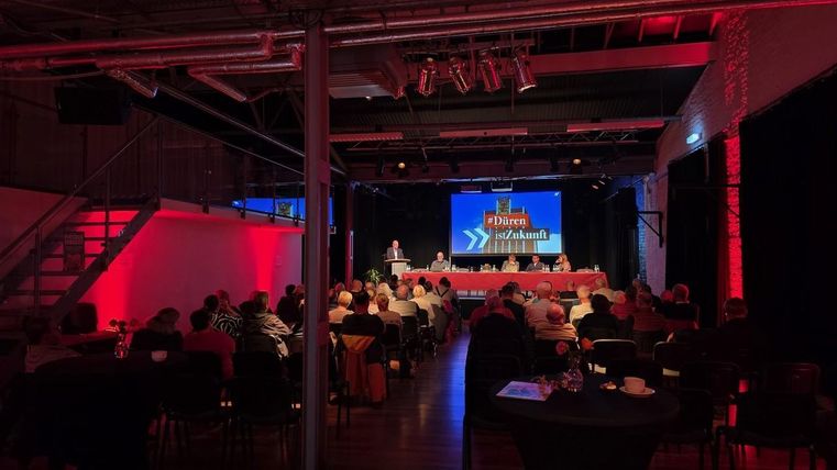 Een conferentie met een spreker voor een publiek in een moderne evenementenruimte. Rode en blauwe verlichting creëren een aantrekkelijke sfeer.