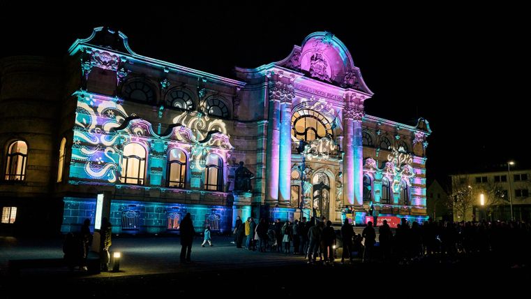 Un bâtiment historique, illuminé de manière colorée la nuit. Beaucoup de gens se tiennent devant et admirent le spectacle lumineux.