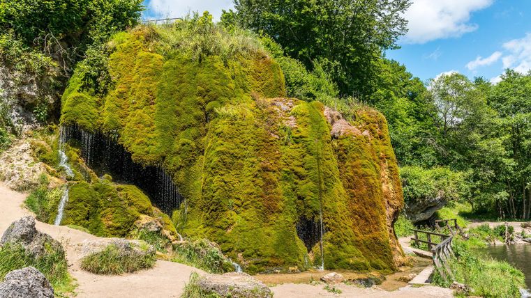 Moosbedeckter Wasserfall bei Nohn, umgeben von üppigem Grün. Ein kleiner Steg führt am Wasser entlang. Der Himmel ist blau mit einigen Wolken.