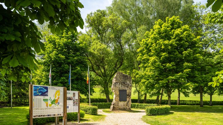 Dreiländereck bei Ouren: Gedenkstein mit Informationstafeln an einem gepflasterten Weg, umgeben von üppig grünem Baumbestand bei sonnigem Wetter.