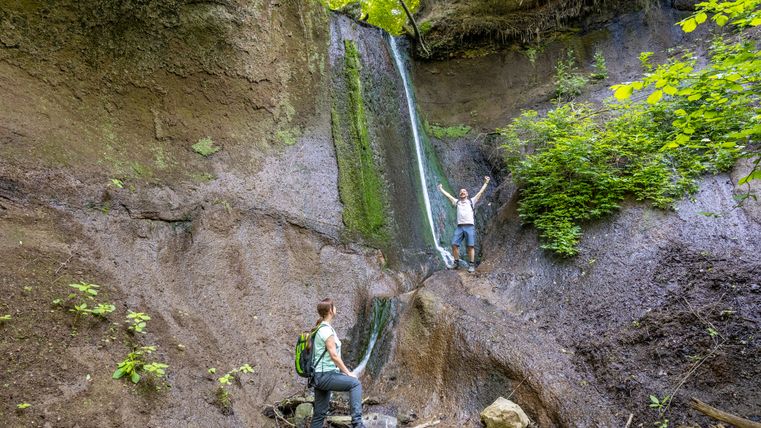 Twee mensen staan bij een kleine waterval in een kloof, omringd door rotsen en planten.
