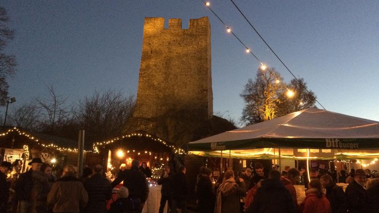 Un marché de Noël avec beaucoup de monde, des stands décorés de façon festive et un vieux château en arrière-plan. L'ambiance est confortable et il fait crépusculaire.