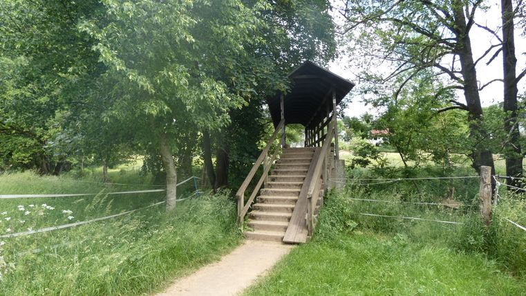Overdekte houten brug over een kleine rivier, omgeven door bomen en weiden.
