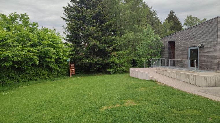 Une pelouse verte avec des arbres et des buissons en arrière-plan. À droite, une vue moderne du bâtiment avec un escalier.