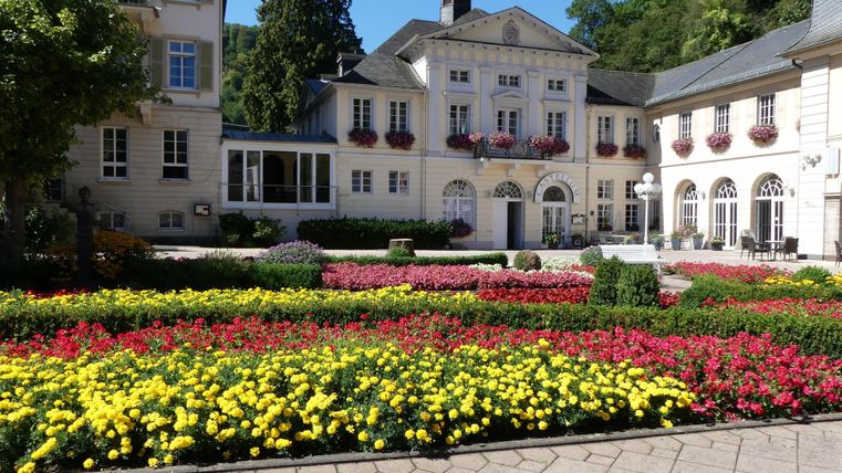 Ein schöner Gartenvorplatz mit bunten Blumenbeeten in Gelb, Rot und Pink. Im Hintergrund ist ein elegantes Gebäude mit dekorativen Fenstern und Pflanzen zu sehen.