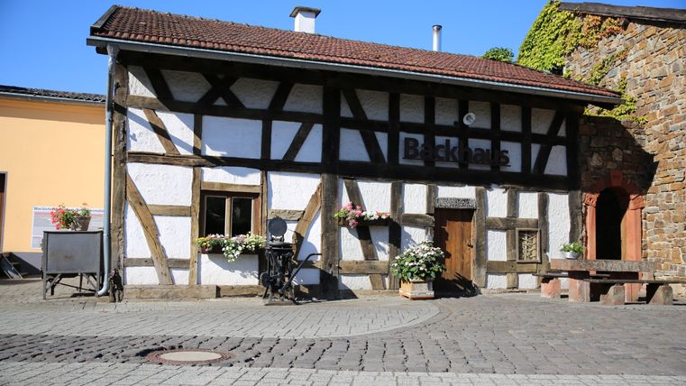 Half-timbered house with the inscription 'Backhaus', surrounded by flowers and a paved square.