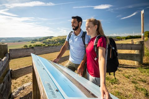 Zwei Personen mit Rucksäcken betrachten eine Informationstafel in einer ländlichen Landschaft.