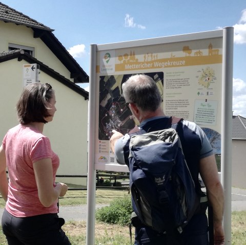 Twee wandelaars staan voor een wandelbord met de tekst 'Metterich wayside crosses' en krijgen informatie over de route. Op de achtergrond zijn huizen te zien., © Tourist-Information Bitburger Land