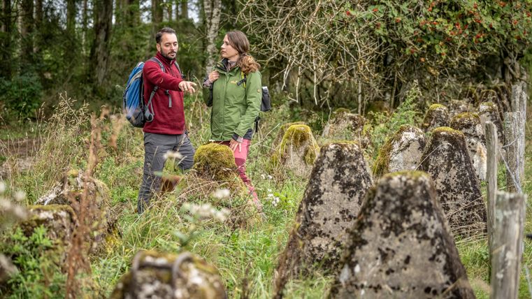 Deux personnes se tiennent entre d'anciens barrages antichars dans une zone boisée.