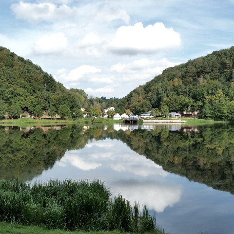 Panorama des Stausee Bitburg mit bewaldeten Hügeln, Gebäuden am Ufer und klarer Spiegelung im Wasser unter blauem Himmel., © TI Bitburger Land