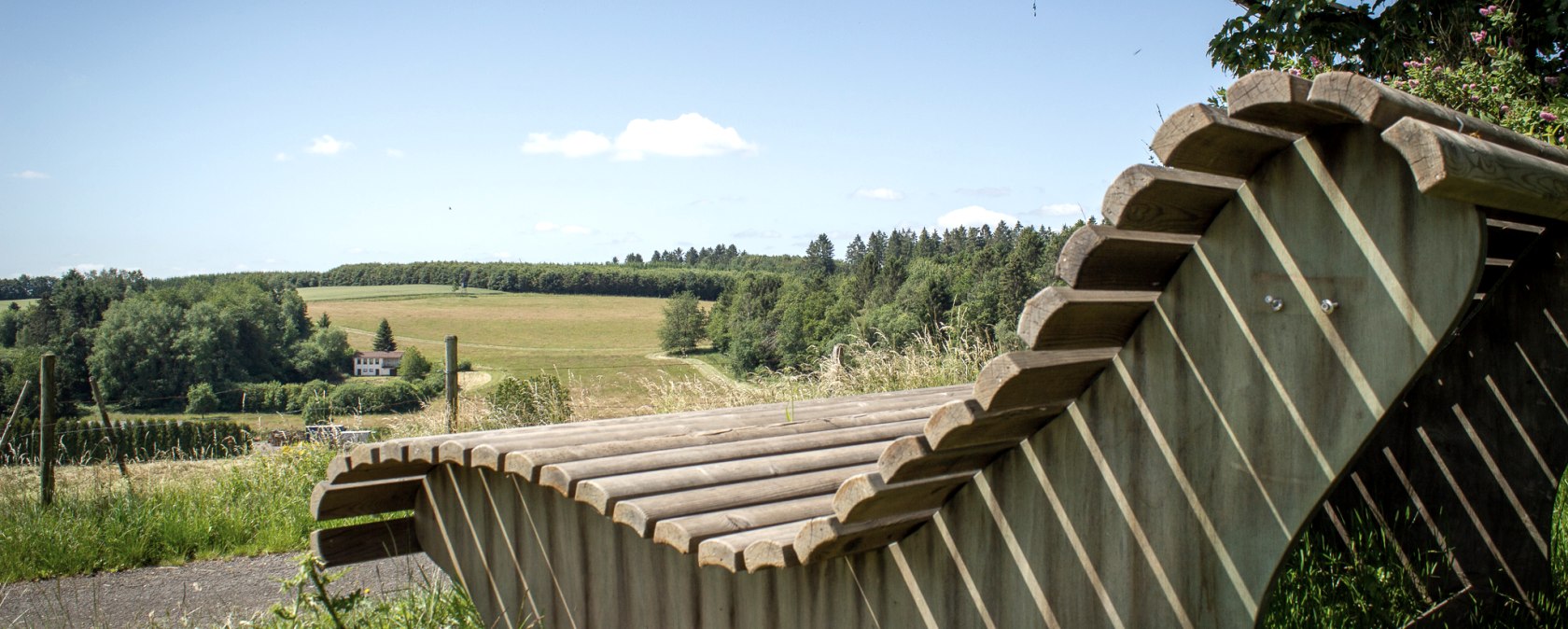 Holzliegebank mit Blick auf grüne Felder und Wälder unter blauem Himmel in Neidenbach., © TI Bitburger Land - Monika Mayer