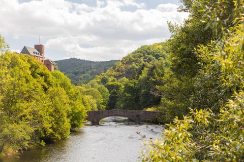 A picturesque landscape with a river and an arch bridge. Surrounded by green trees and gentle hills under a cloudy sky.