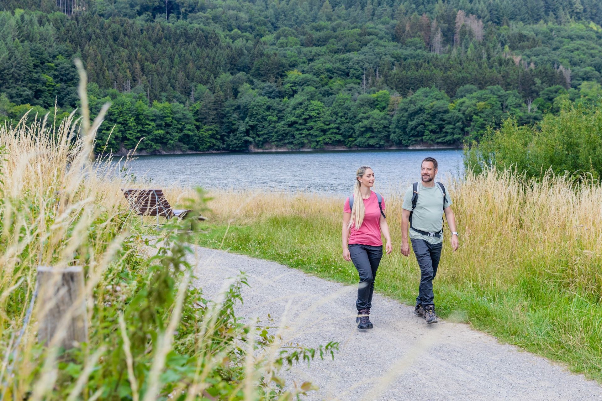 Zwei Wanderer gehen auf einem Schotterweg am Ufer des Rursees entlang. Umgeben von hohen Gräsern und bewaldeten Hügeln im Hintergrund genießen sie die Natur an einem sommerlichen Tag.