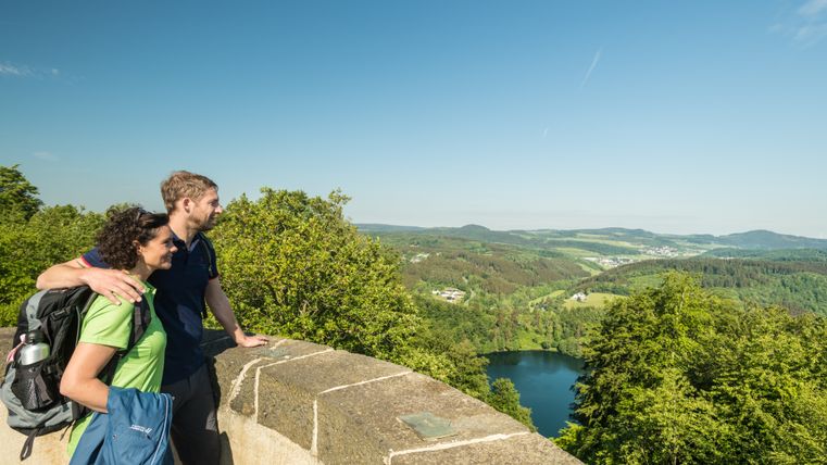 Two people stand on a vantage point and look out over a green landscape with a lake.